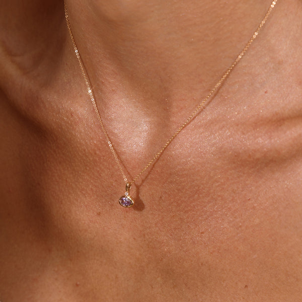 A close up lifestyle photo of a person's chest wearing a dainty gold chain pendant necklace with a gold oval charm featuring an oval bezel-set light purple gemstone.