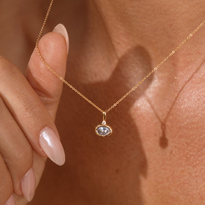 A close up lifestyle photo of a person's neck wearing a dainty gold chain necklace with a gold oval charm featuring a light blue oval gemstone, being held by a hand to showcase the pendant.
