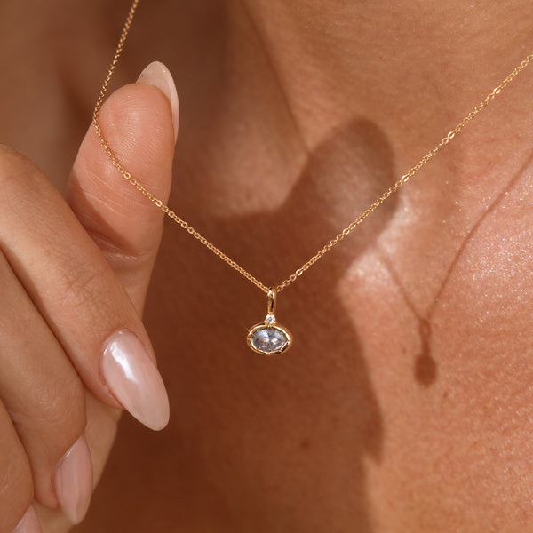 A close up lifestyle photo of a person's neck wearing a dainty gold chain necklace with a gold oval charm featuring a light blue oval gemstone, being held by a hand to showcase the pendant.
