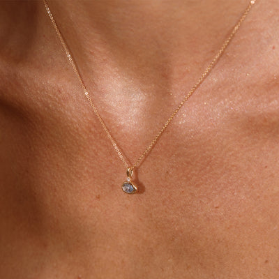 A close up lifestyle photo of a person's chest wearing a dainty gold chain pendant necklace with a gold oval charm featuring an oval bezel-set light blue gemstone.