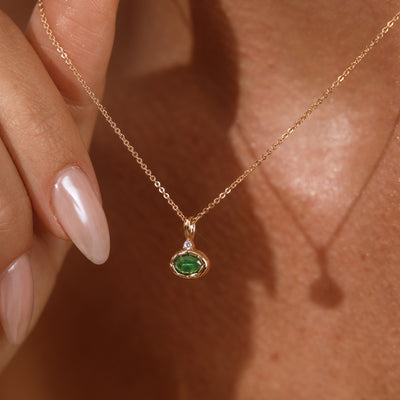 A close up lifestyle photo of a person's neck wearing a dainty gold chain necklace with a gold oval charm featuring a dark green oval gemstone, being held by a hand to showcase the pendant.