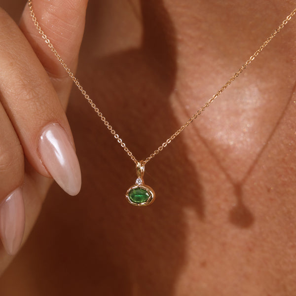 A close up lifestyle photo of a person's neck wearing a dainty gold chain necklace with a gold oval charm featuring a dark green oval gemstone, being held by a hand to showcase the pendant.