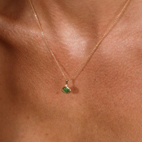 A close up lifestyle photo of a person's chest wearing a dainty gold chain pendant necklace with a gold oval charm featuring an oval bezel-set green gemstone.