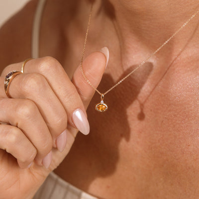 A close up lifestyle photo of a person's neck wearing a dainty gold chain necklace with a gold oval charm featuring an amber oval gemstone, being held by one hand.