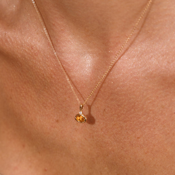 A close up lifestyle photo of a person's chest wearing a dainty gold chain pendant necklace with a gold oval charm featuring an oval bezel-set amber gemstone.