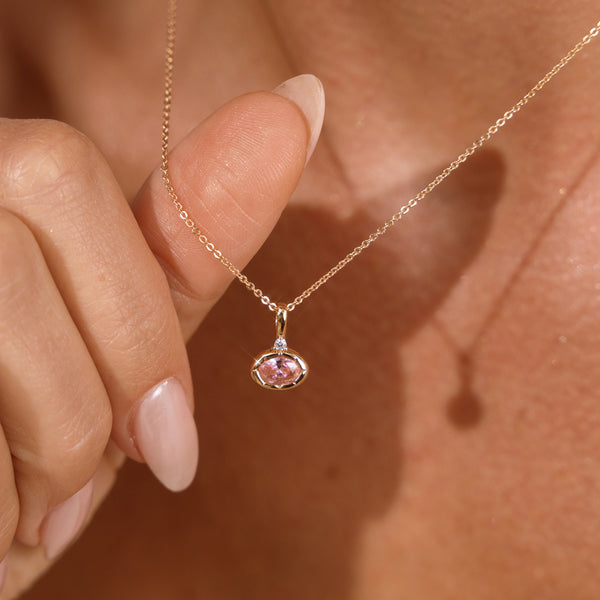 A close up lifestyle photo of a person's neck wearing a dainty gold chain necklace with a gold oval charm featuring a pink oval gemstone, being held by one hand.