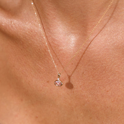 A close up lifestyle photo of a person's chest wearing a dainty gold chain pendant necklace with a gold oval charm featuring an oval bezel-set pink gemstone.
