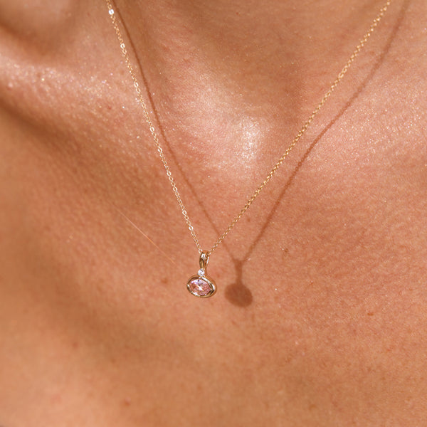 A close up lifestyle photo of a person's chest wearing a dainty gold chain pendant necklace with a gold oval charm featuring an oval bezel-set pink gemstone.