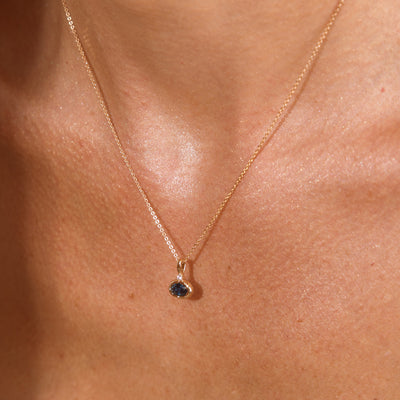 A close up lifestyle photo of a person's chest wearing a dainty gold chain pendant necklace with a gold oval charm featuring an oval bezel-set blue-green gemstone.