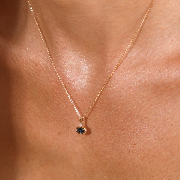 A close up lifestyle photo of a person's chest wearing a dainty gold chain pendant necklace with a gold oval charm featuring an oval bezel-set blue-green gemstone.
