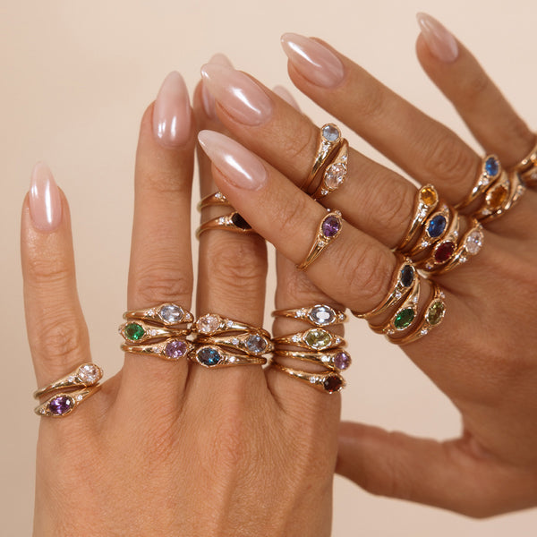 A close up lifestyle shot of a person's hands with stacks of multiple gold signet rings, all with different colored oval bezel-set gemstones, against a beige background.