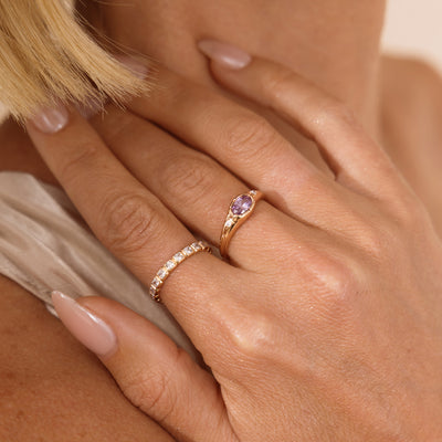 A close up lifestyle photo of a hand wearing two rings, one gold oval signet ring featuring a light purple oval bezel-set gemstone, and a gold eternity style ring with square clear bezel-set stones.