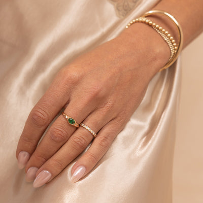 A close up lifestyle shot of a hand with a gold oval signet ring, featuring a green bezel-set oval gemstone with two clear accent stones, paired next to a gold eternity ring with clear, square, bezel-set stones and a variety of gold bracelets.