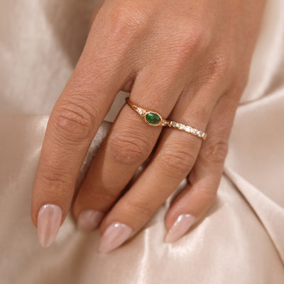 A close up lifestyle shot of a hand with a gold oval signet ring, featuring a green bezel-set oval gemstone with two clear accent stones, paired next to a gold eternity ring with clear, square, bezel-set stones.