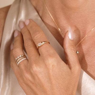 A close up lifestyle shot of a hand against a chest with a gold oval signet ring featuring a clear oval, bezel-set gemstone, and a stack of different gold rings with gemstones and pearls.