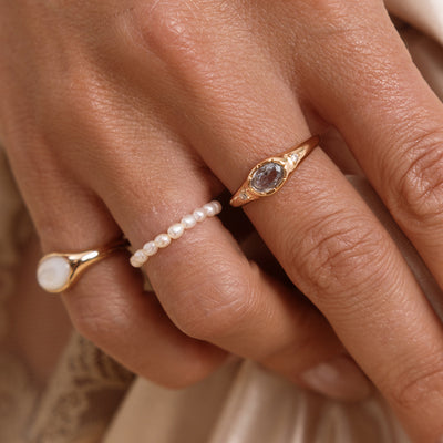 A close up lifestyle shot of a hand with a gold oval signet ring, featuring a light blue bezel-set oval gemstone with two clear accent stones, paired next to a gold signet ring with mother of pearl inlay and a pearl eternity ring.