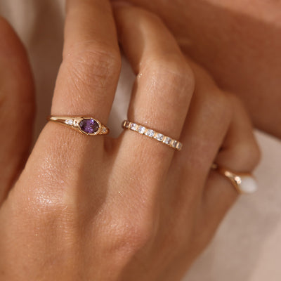 A close up lifestyle shot of a hand wearing three rings, one gold signet ring with a purple bezel-set oval gemstone, one gold eternity style ring with clear, square, bezel-set stones, and a gold signet ring with a mother of pearl inlay.