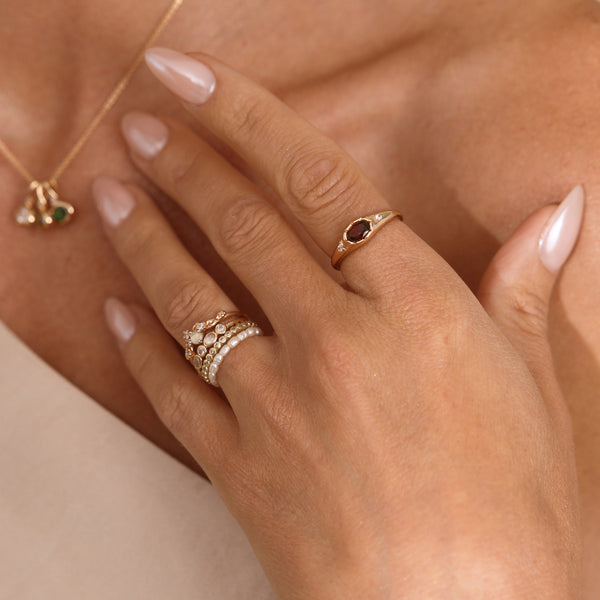 A close up lifestyle shot of a hand featuring a gold oval gemstone signet ring with a red stone, and a stack of multiple gold rings and a pearl eternity ring.