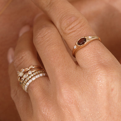 A close up lifestyle shot of a hand featuring a gold oval gemstone signet ring with a red stone, and a stack of multiple gold rings and a pearl eternity ring.