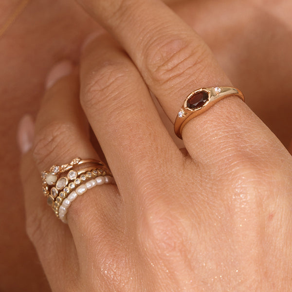 A close up lifestyle shot of a hand featuring a gold oval gemstone signet ring with a red stone, and a stack of multiple gold rings and a pearl eternity ring.