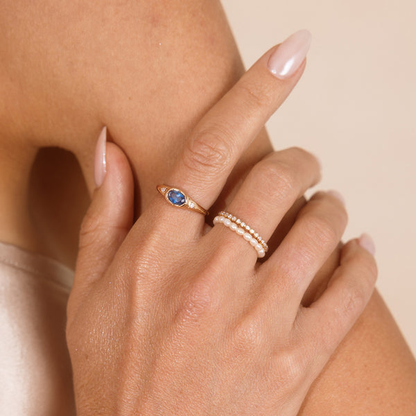 A close up lifestyle shot of a hand featuring a gold oval gemstone signet ring with a blue stone, and a stack of two eternity rings, one with round clear stones and the other pearls.