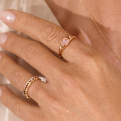 A close up lifestyle photo of a hand wearing a gold signet ring with a pink bezel-set oval gemstone, and two eternity style rings with round clear stones stacked on another finger.