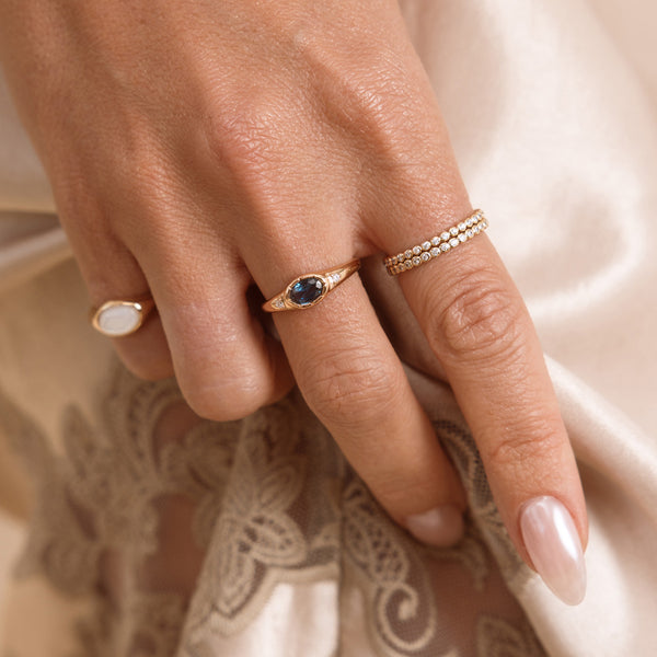 A close up lifestyle shot of a hand featuring a gold oval gemstone signet ring with a blue-green stone, a stack of two gold eternity rings with round clear stones and a gold signet ring with mother of pearl inlay.
