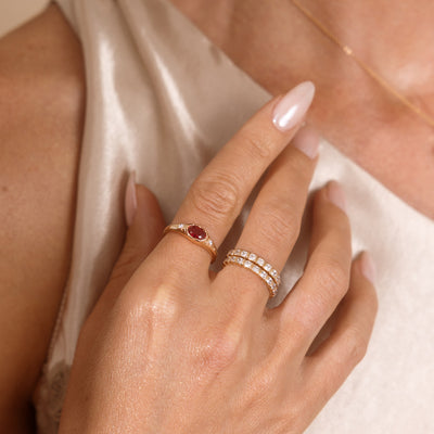 A close up lifestyle shot of a hand featuring a gold oval gemstone signet ring with a red stone, and a stack of two gold eternity rings with clear, square, bezel-set stones.