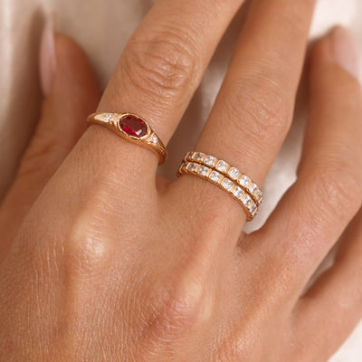 A close up lifestyle shot of a hand featuring a gold oval gemstone signet ring with a red stone, and a stack of two gold eternity rings with clear, square, bezel-set stones.