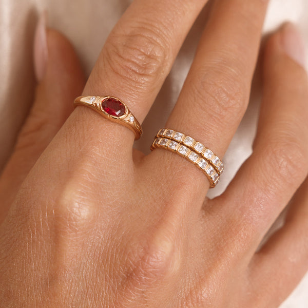 A close up lifestyle shot of a hand featuring a gold oval gemstone signet ring with a red stone, and a stack of two gold eternity rings with clear, square, bezel-set stones.