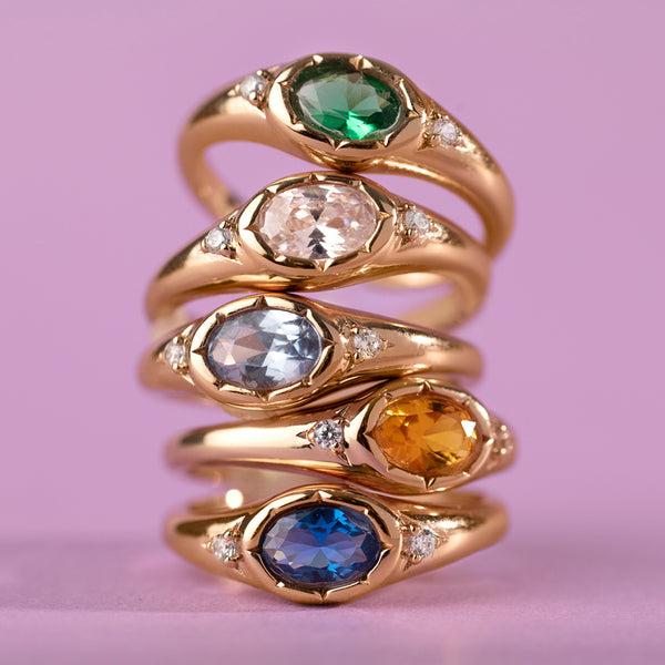 A stack of gold oval signet rings, featuring a green, clear, light blue, amber, and dark blue oval bezel-set gemstone with two clear accent stones on either side, shown against a pink background.