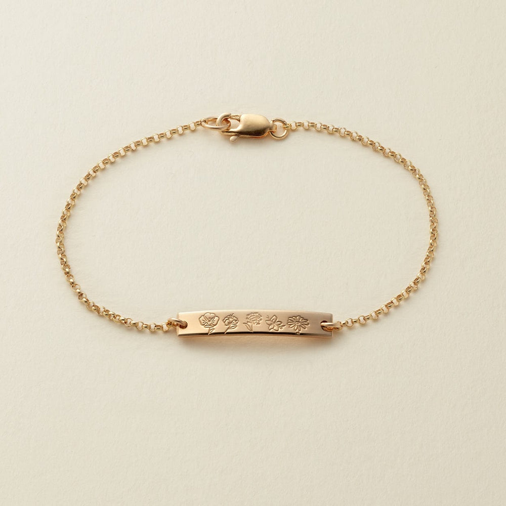 A delicate gold chain bracelet with a central bar engraved with different flower designs, displayed on a light beige textured background.