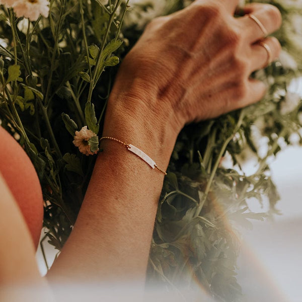 A person holding a bouquet of greenery with white flowers, wearing a delicate gold chain bracelet with a central bar engraved with different flower designs.