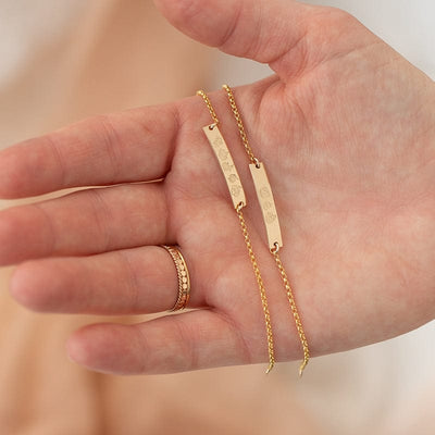 A person's hand holding two delicate gold chain bracelets with a central bar engraved with different flower designs, with a stack of different gold rings on their finger.