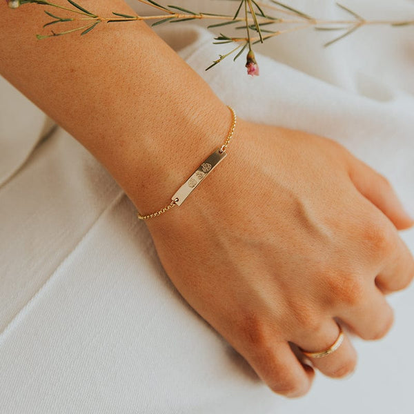 A person's wrist resting on a white linen surface with a thin sprig of greenery, showcasing a delicate gold chain bracelet with a central bar engraved with different flower designs.