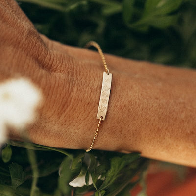 A person's wrist in front of green foliage wearing a delicate gold chain bracelet with a central bar engraved with different flower designs.