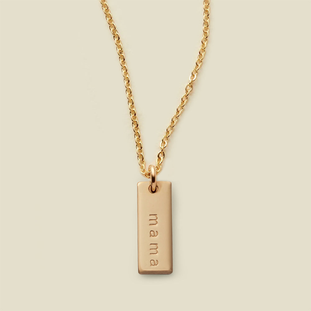 A gold chain necklace with a rectangular gold bar pendant engraved with the word 'mama', shown against a solid light beige background.