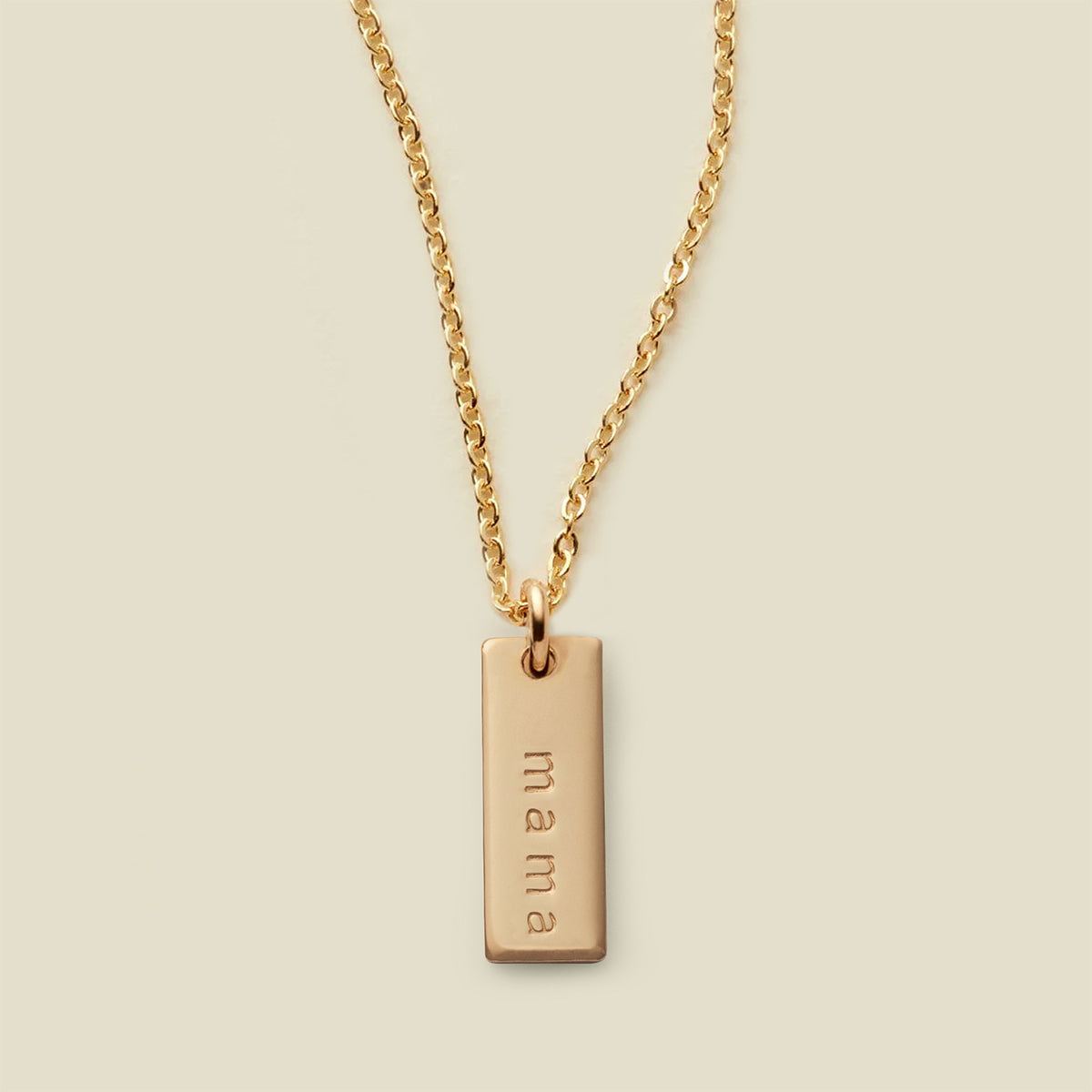 A gold chain necklace with a rectangular gold bar pendant engraved with the word 'mama', shown against a solid light beige background.