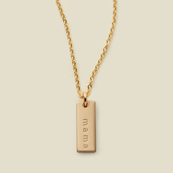 A gold chain necklace with a rectangular gold bar pendant engraved with the word 'mama', shown against a solid light beige background.