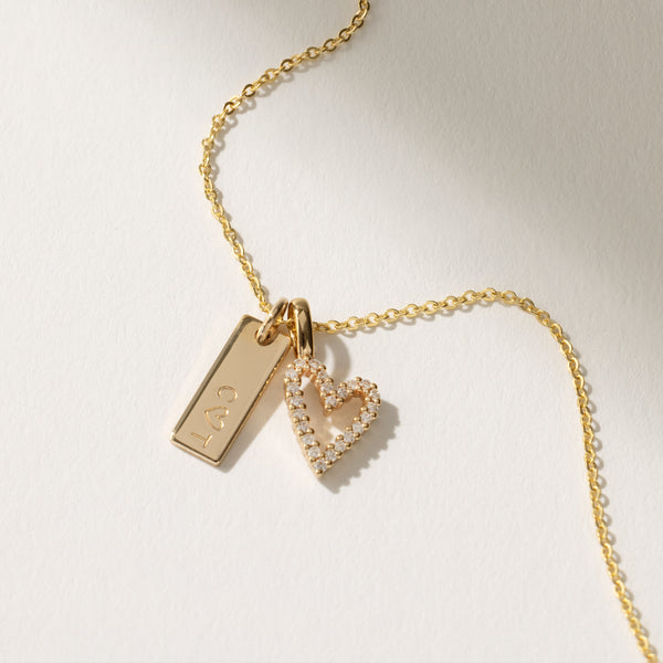 A delicate gold chain necklace with a gold heart charm covered in clear accent stones and a rectangular bar charm engraved with the letters 'C ♡ T'.