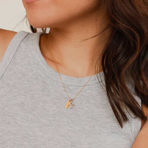 A close-up of a person in a ribbed high neck grey tank top, wearing a delicate gold chain necklace with a heart charm and a small rectangular bar charm.