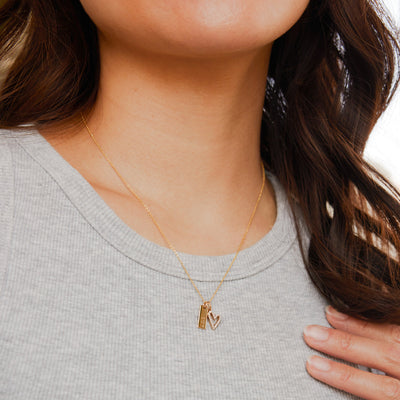 A close-up of a person in a ribbed high neck grey tank top, wearing a delicate gold chain necklace with a heart charm and a small rectangular bar charm.