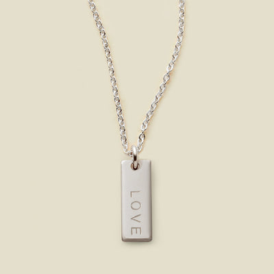 A silver chain necklace with a rectangular silver bar pendant engraved with the word 'LOVE', shown against a solid light beige background.