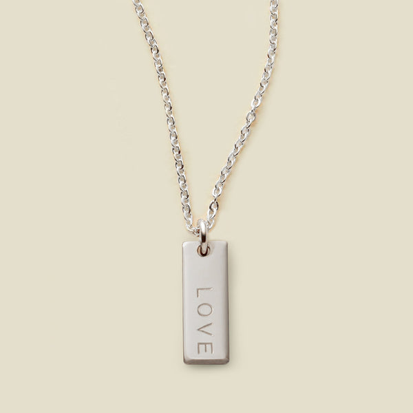 A silver chain necklace with a rectangular silver bar pendant engraved with the word 'LOVE', shown against a solid light beige background.