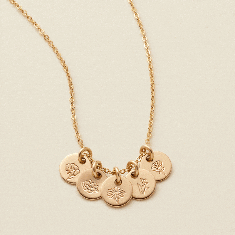 A gif of a delicate gold chain necklace with five small round flat discs, each with their own floral design engraved. The floral designs on different discs change every few seconds.
