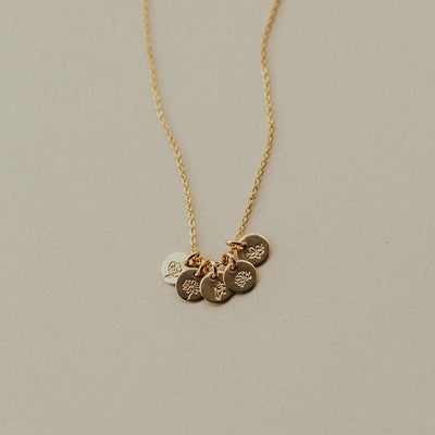 A dainty gold pendant chain necklace with five small round flat discs, each engraved with a different birth flower design and shown on a solid beige background.
