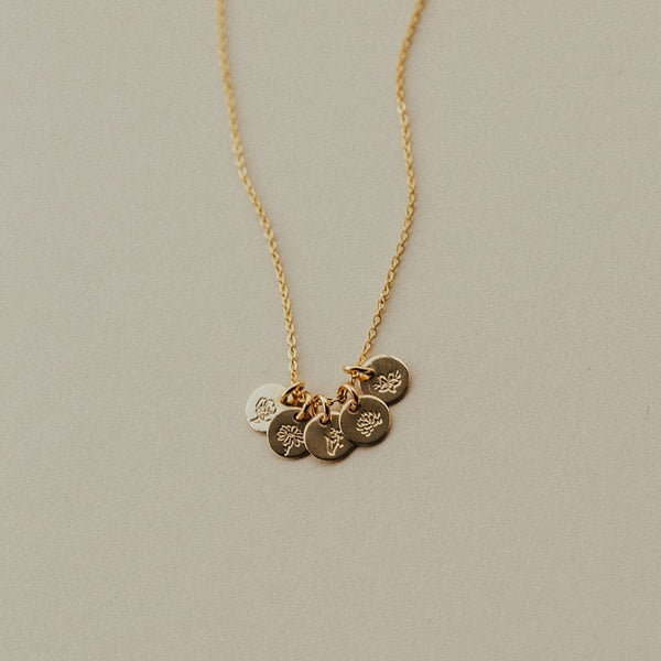 A dainty gold pendant chain necklace with five small round flat discs, each engraved with a different birth flower design and shown on a solid beige background.