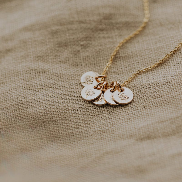 A delicate gold chain necklace with five small round discs, each engraved with a different floral design and shown on a beige linen surface.