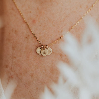 A close-up of a person's chest wearing a gold pendant chain with three small round flat discs, each engraved with a different floral design, with white foliage blurred in the foreground.