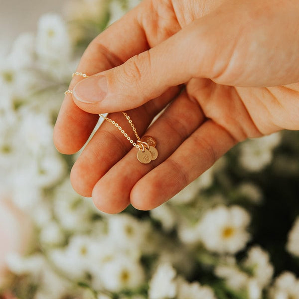 A hand holding a gold chain necklace with three small round flat discs, each with a different flower design, with a bouquet of white flowers blurred in the background.